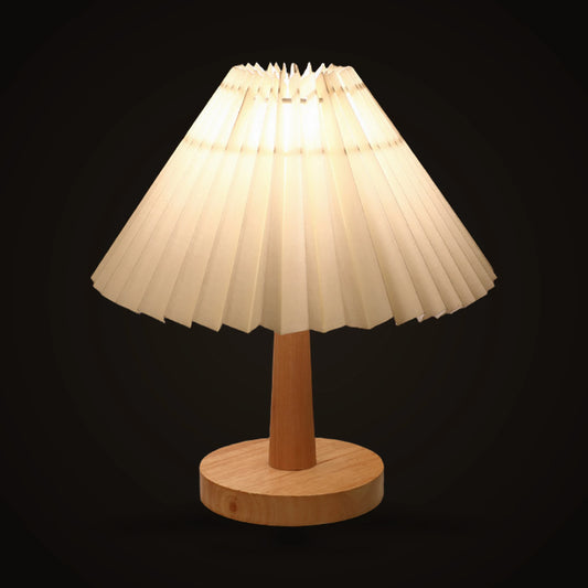 The Lumivano Pleat on the dark brand ground, switched on, the warm light reading through the off-white pleats as alternating bands of cream and shadow above the pale wooden stem and round foot.