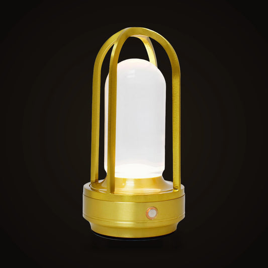 The Lumivano Arbour on the dark brand ground: a small gold aluminium lantern lamp with slender ribs rising from a round base and meeting in an arch over a glowing milky white core, with a small round button on the front of the base.
