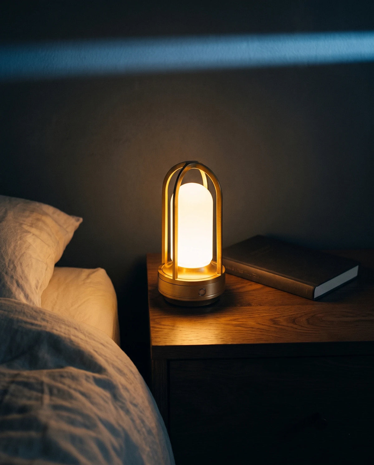 The Lumivano Arbour glowing on a dark oak bedside table beside the edge of a bed made with ivory linen, a small circle of warm light on the wood and a closed book in shadow against a dark plaster wall, with a cool band of moonlight across the top of the wall.