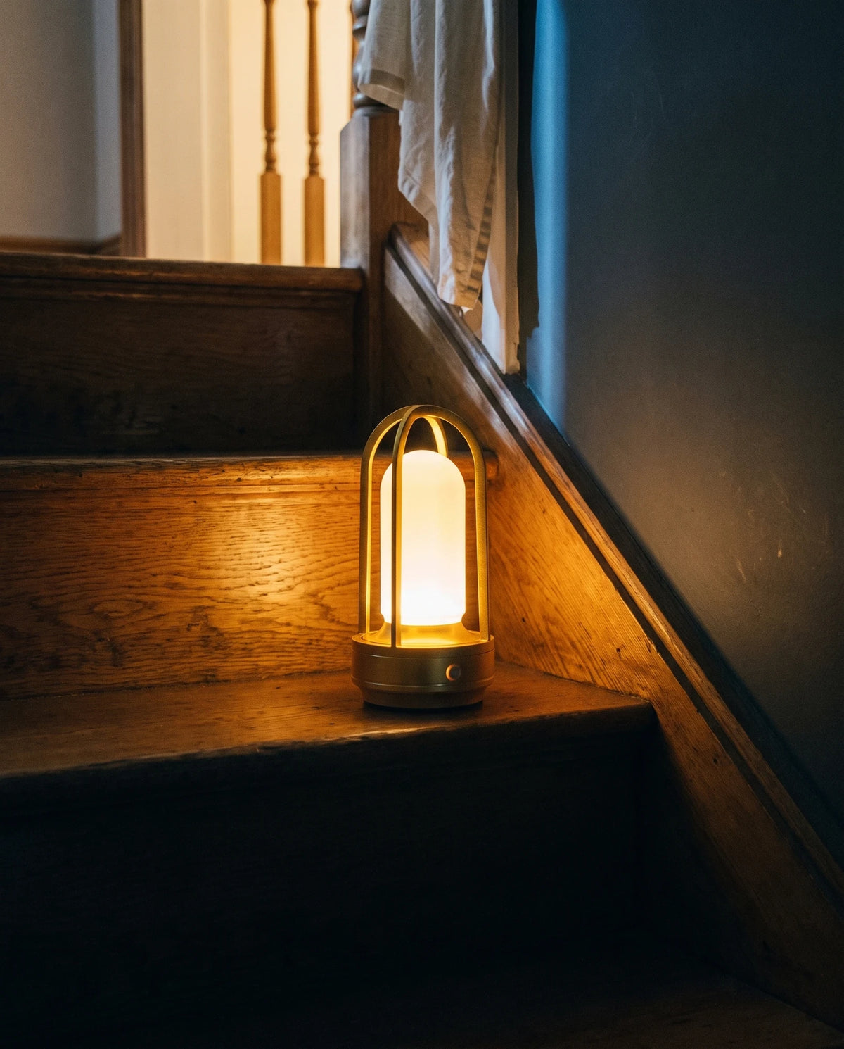 The Lumivano Arbour standing alone on a worn oak stair tread with no cable attached, its warm glow lighting the grain of the step, with turned wooden balusters and an ivory linen throw over the banister above and a cool blue light along the dark wall to the right.