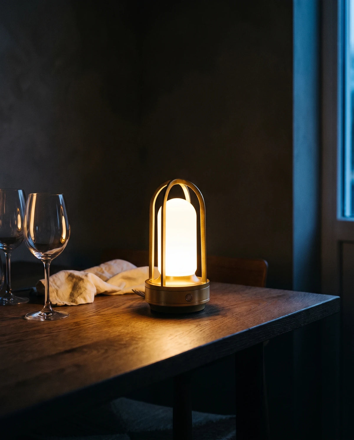The Lumivano Arbour near the end of a dark oak dining table after dinner, its warm light spreading over a crumpled ivory linen napkin and two empty wine glasses, with a dark plaster wall behind and a strip of cool dusk at a window on the right.
