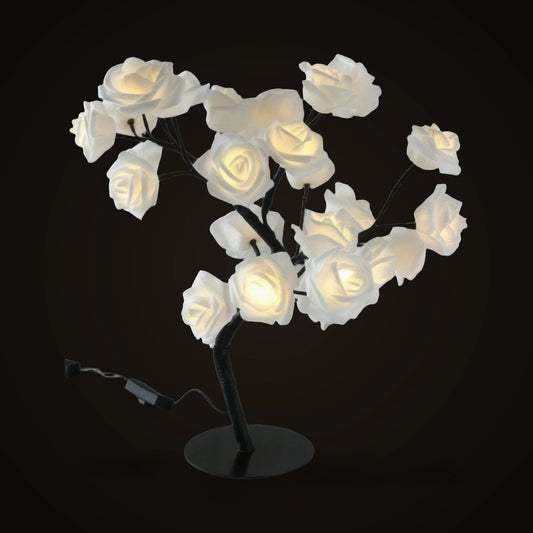 The Lumivano Blossom on the dark brand ground: a small tree of black branches carrying cream-white roses that glow warmly from inside, on a round black base, with its attached black cable and inline on/off switch lying beside it.