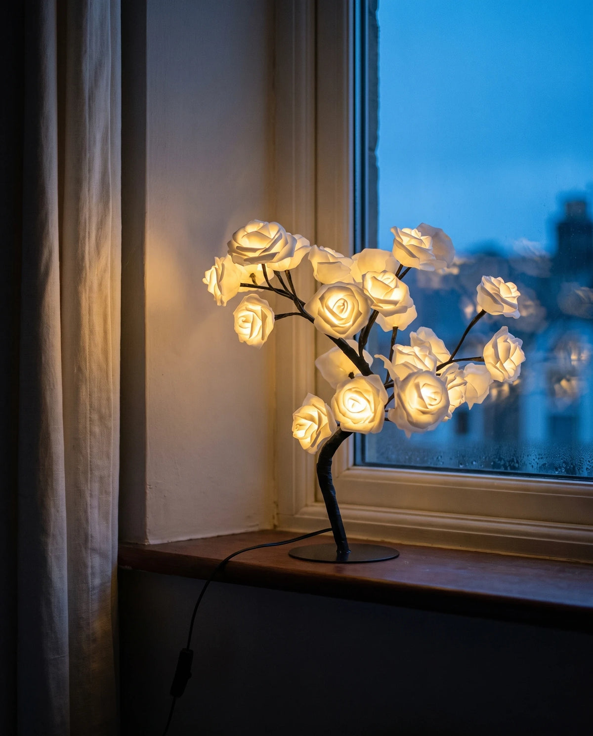 The Lumivano Blossom lit on a painted windowsill at dusk, its cream-white roses glowing against the deep blue sky beyond the glass, with the black cable and its inline on/off switch trailing across the sill.