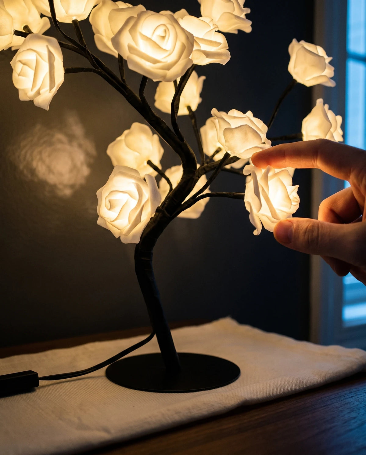 Two fingertips moving one of the Lumivano Blossom's slender black wire branches to reposition a cream-white rose, the lit roses throwing a dappled warm pattern across the dark wall behind and the black cable running from the base to the left.