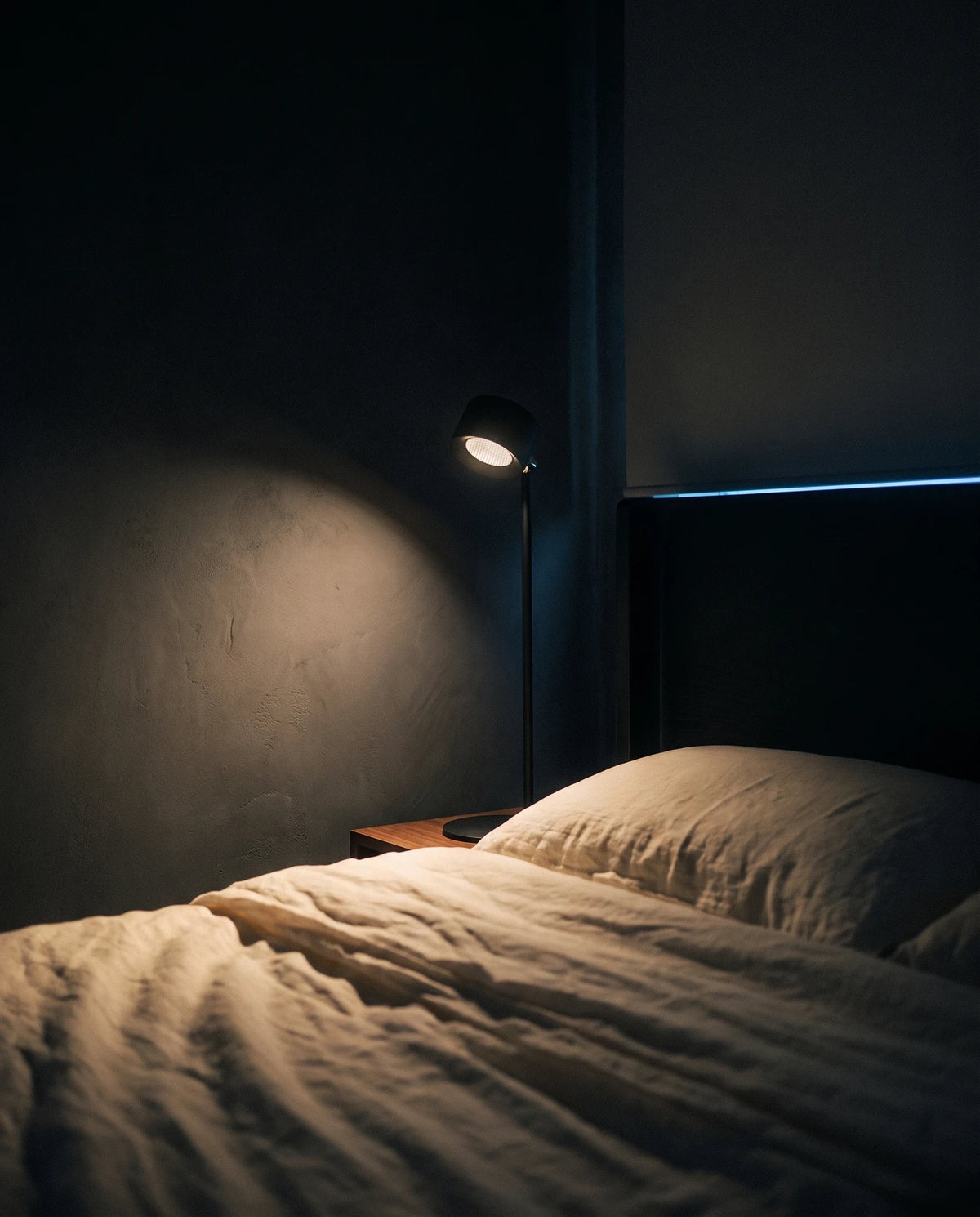 The Lumivano Halo on a walnut bedside table with its head turned to the wall, throwing a soft warm oval of light across dark plaster, with rumpled ivory linen bedding below and a thin cool line of moonlight along the headboard.
