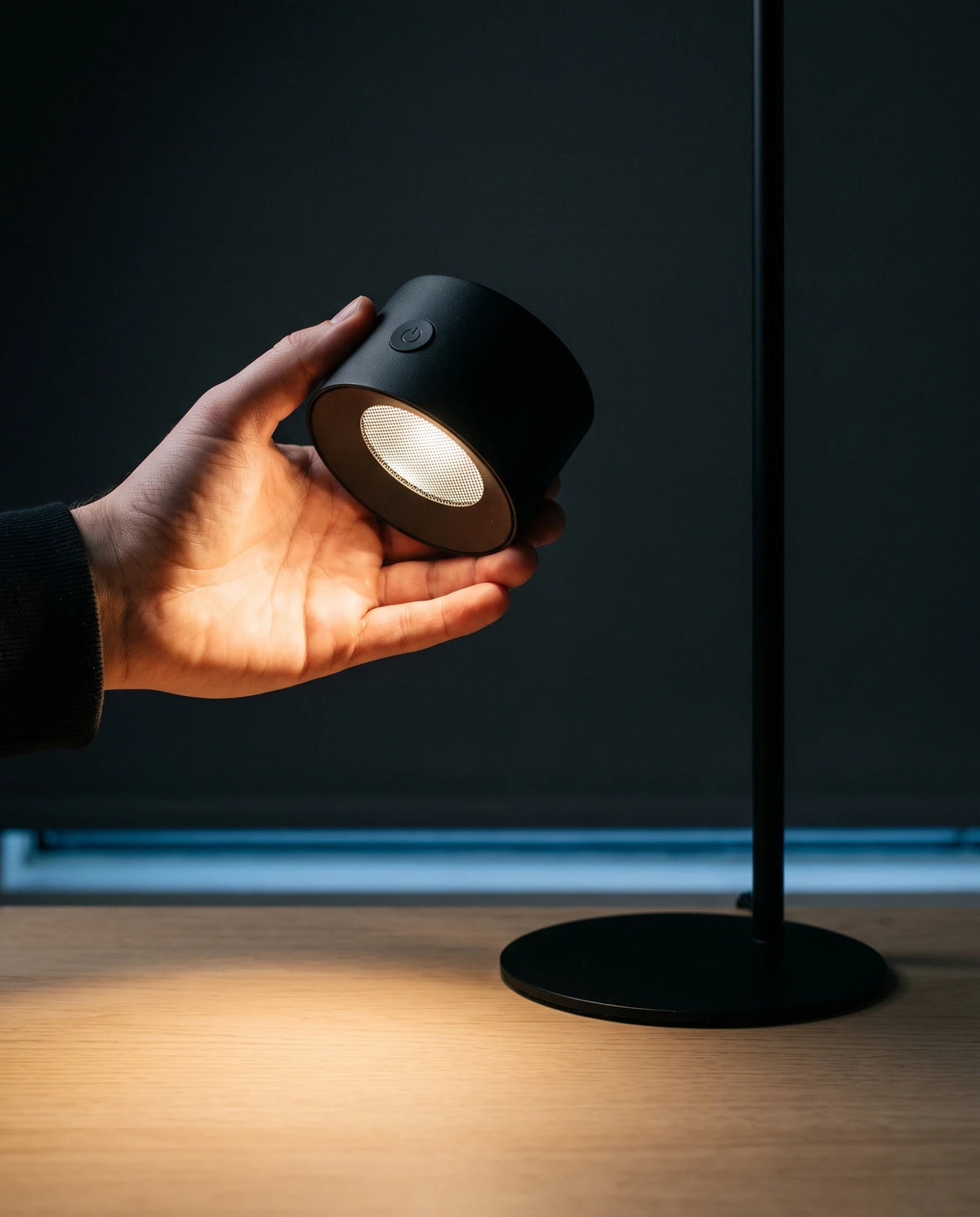 A hand lifting the Lumivano Halo's cylindrical head off its stem, the head glowing warm in the palm with its mesh lens facing down, the stem and its round black base standing on the pale oak desk alongside.
