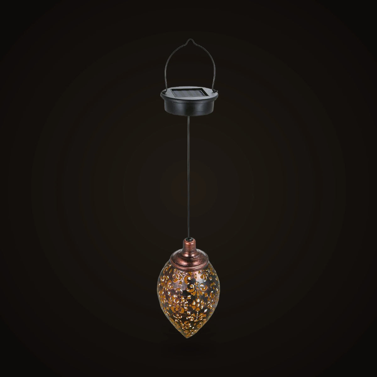 The hanging solar lantern switched off, on the dark brand ground: a round black cap with a small square solar panel on top and a thin black wire handle arching over it, a thin black cord running straight down from the cap, and below it a teardrop-shaped metal lantern with a copper-toned collar and neck, its dark shell pierced all over with small scrolls and flowers.