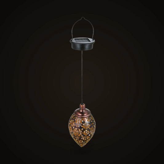 The hanging solar lantern switched off, on the dark brand ground: a round black cap with a small square solar panel on top and a thin black wire handle arching over it, a thin black cord running straight down from the cap, and below it a teardrop-shaped metal lantern with a copper-toned collar and neck, its dark shell pierced all over with small scrolls and flowers.