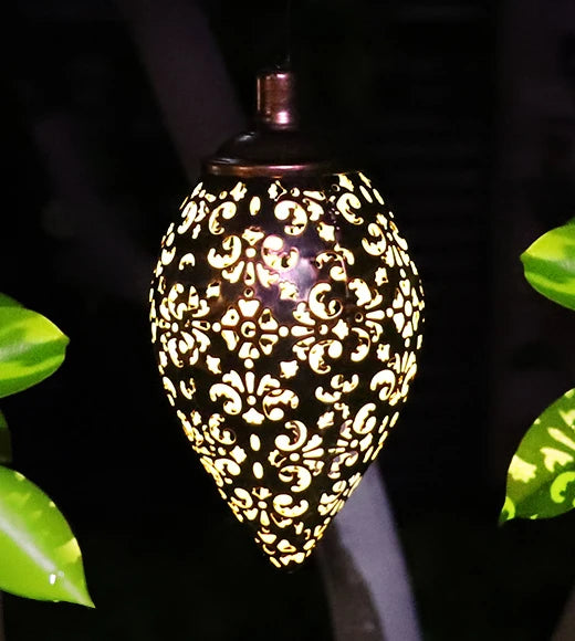 A close view of the lit lantern hanging among dark leaves: the copper-toned collar and neck at the top and the teardrop shell below, cut through with a dense pattern of four-petal flowers, paired scrolls and small teardrop openings, all glowing warm white against the dark metal.