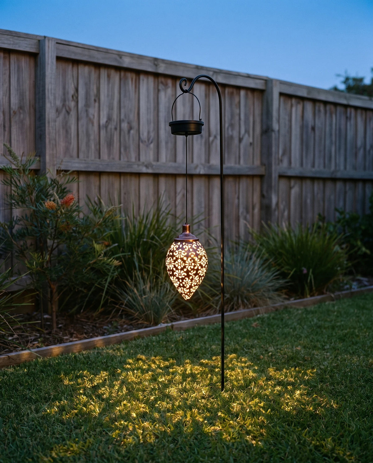 The lantern lit at dusk in a small back garden, hanging from a slim black metal garden stand pushed into the lawn, its black solar cap at the top and the glowing pierced teardrop below, throwing a scatter of small warm light spots onto the grass, with a garden bed and a timber fence behind. The stand is not included.