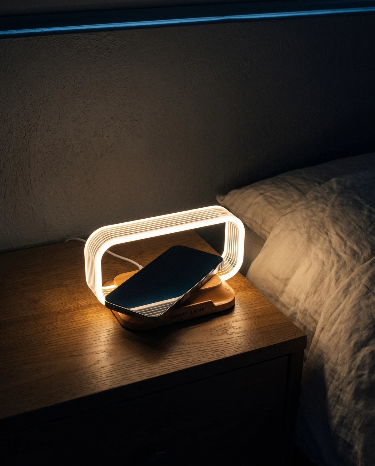 The Lumivano Lintel on a dark oak nightstand beside an unmade bed at night: a small lamp whose hoop of clear acrylic glows warm white over a low wood-look base, a smartphone lying diagonally on the charging pad and reaching almost from one side of the hoop to the other, with a thin white cable running away behind it and a line of cool moonlight along the top of the wall.