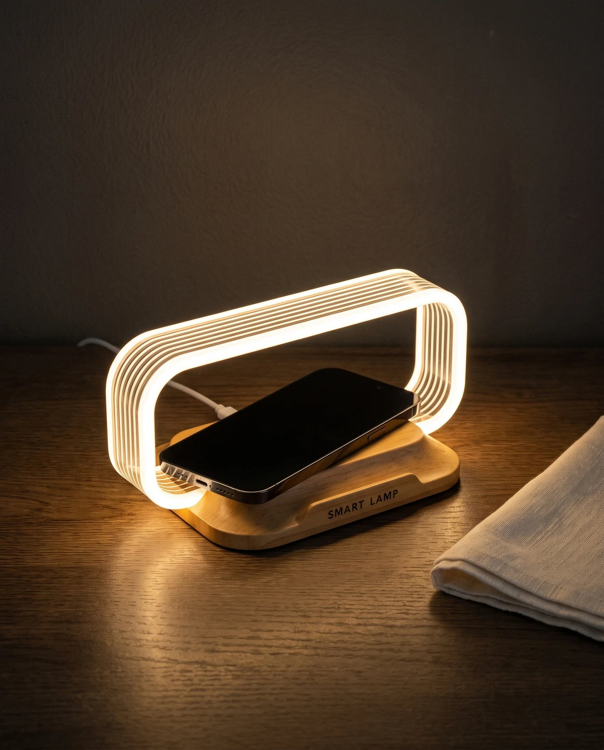 The Lumivano Lintel on a dark oak desk in the evening next to a folded linen cloth: a smartphone lies across the wood-look charging pad, as long as the glowing acrylic hoop is wide, with a white USB cable leaving the back of the base.