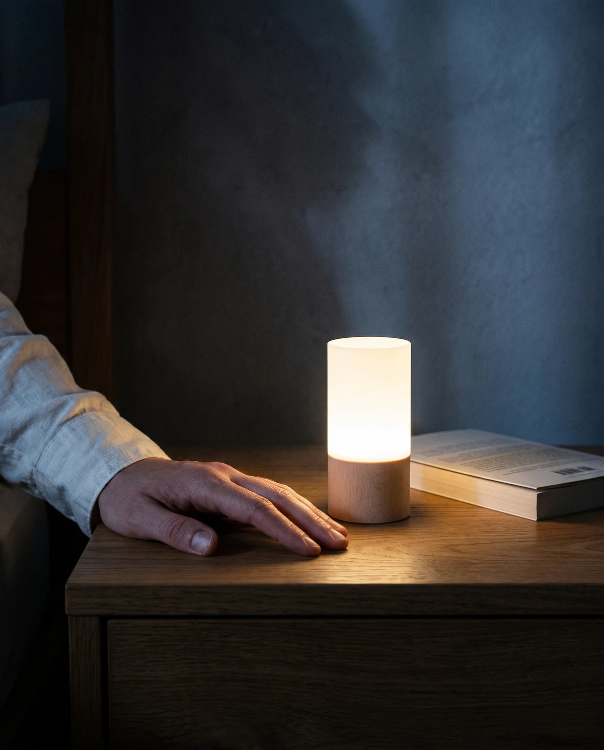 An adult hand resting flat on a dark oak bedside table right beside the glowing night light, which is about as tall as the hand is long from the wrist to the knuckles, with a closed paperback lying behind it.
