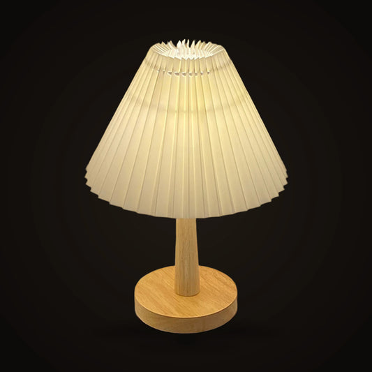 The Lumivano Pleat on the dark brand ground: a wide cone of off-white pleated fabric gathered into sharp vertical folds, lit from inside, on a tapered pale wooden stem and a round wooden foot.
