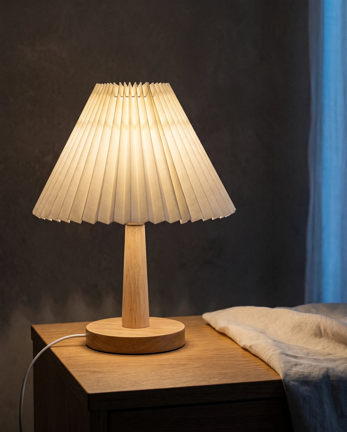 The Lumivano Pleat lit on an oak bedside table against a dark plaster wall, warm light reading through every pleat of the off-white shade, with its white USB lead running visibly across the oak and out of frame and a cool blue curtain at the edge.