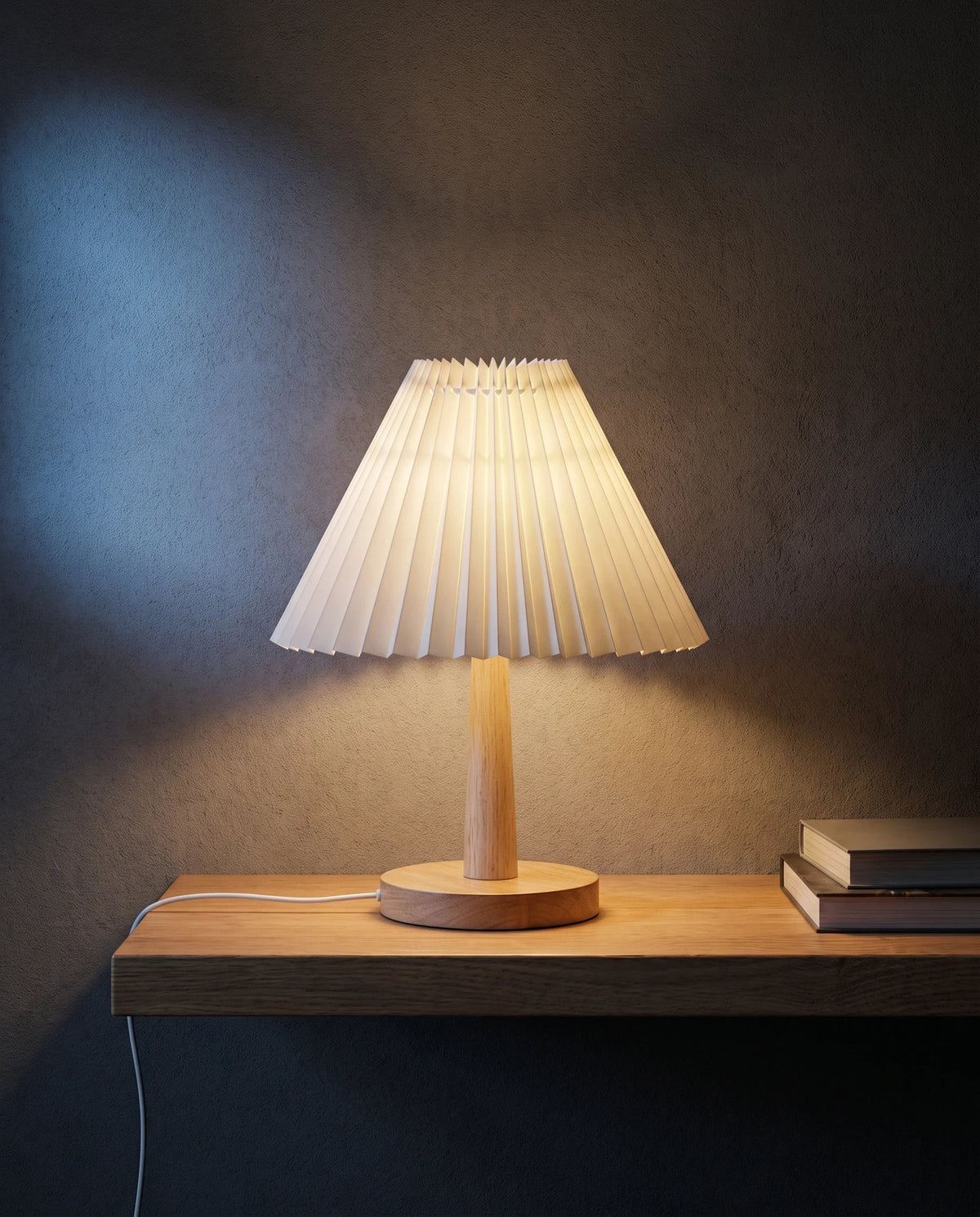 The Lumivano Pleat on a floating oak shelf against a dark plaster wall, its warm light spilling wide across the shelf and up the plaster, with the white USB lead lying along the shelf and dropping over the front edge and two closed books in shadow.