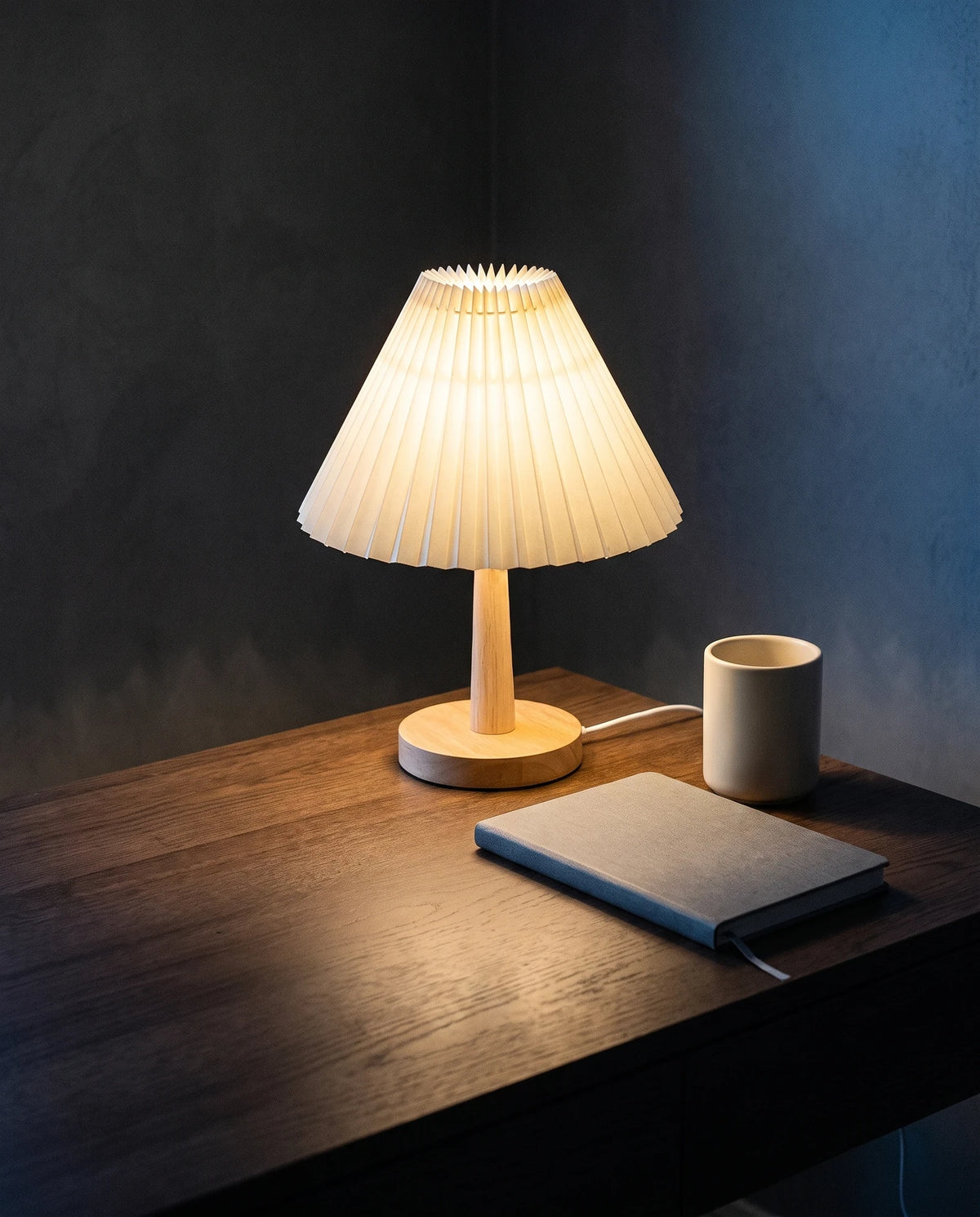 The Lumivano Pleat lit at the back corner of a dark oak writing desk against a dark plaster wall, its warm light spreading across the desktop past a closed notebook and a plain ceramic cup, with its white USB lead running behind the notebook and over the back edge.