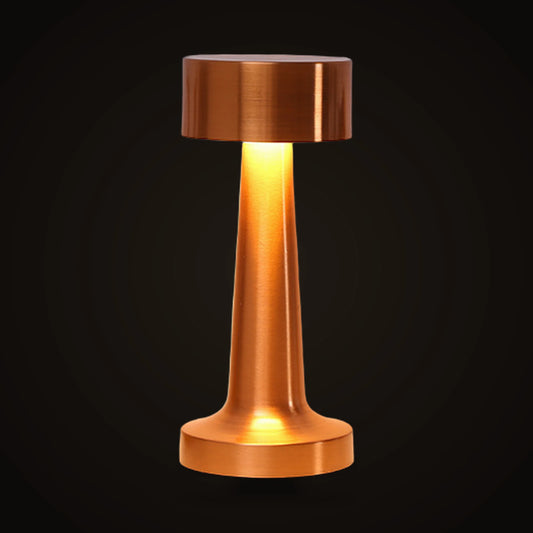 The Lumivano Tavern on the dark brand ground: a small rose gold metal table lamp with a flat circular cap, a slender tapered column and a low round foot, lit so a warm glow escapes from under the cap and runs down the column.