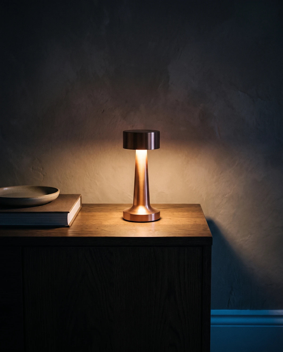 The Lumivano Tavern on a dark oak sideboard, throwing a warm ring of light onto the wood and a soft gradient up a dark plaster wall, with a closed book and a shallow ceramic dish left in shadow and a cool blue line of moonlight along the skirting.