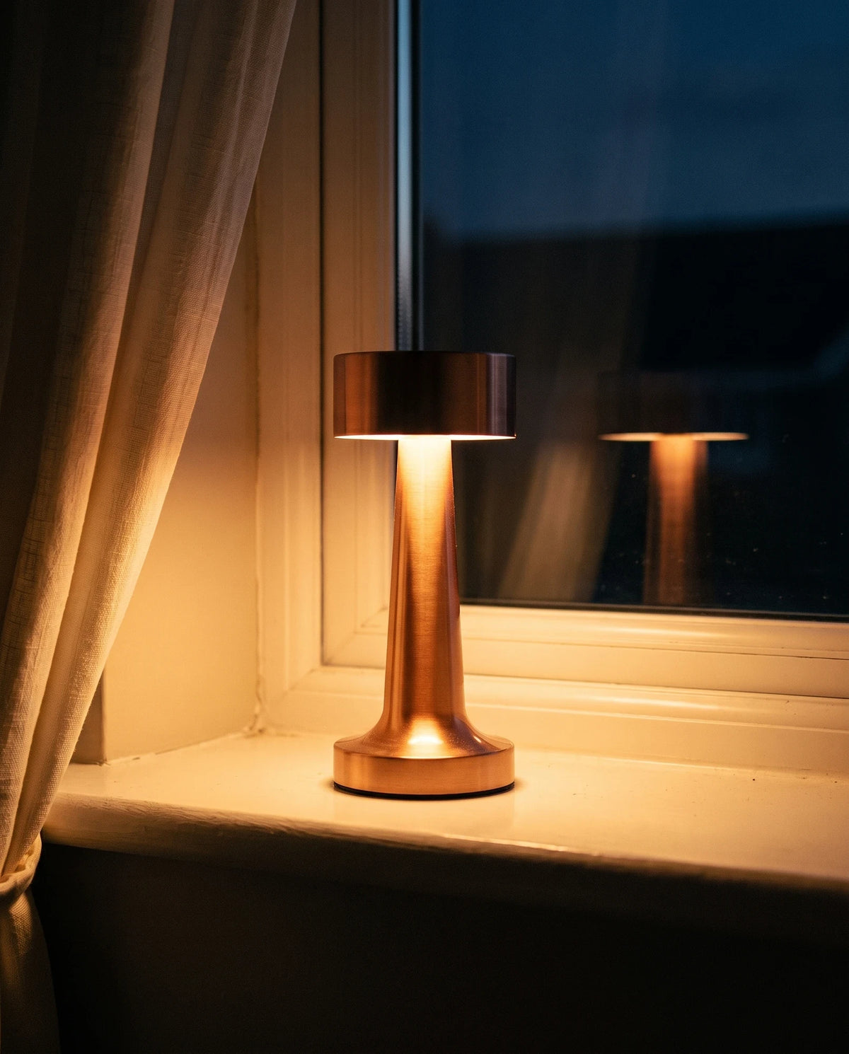 The Lumivano Tavern alone on a painted windowsill at dusk with no cable attached, its warm glow falling on the sill paint and reflecting faintly in the dark glass, with an ivory linen curtain at the left and deep blue evening outside.
