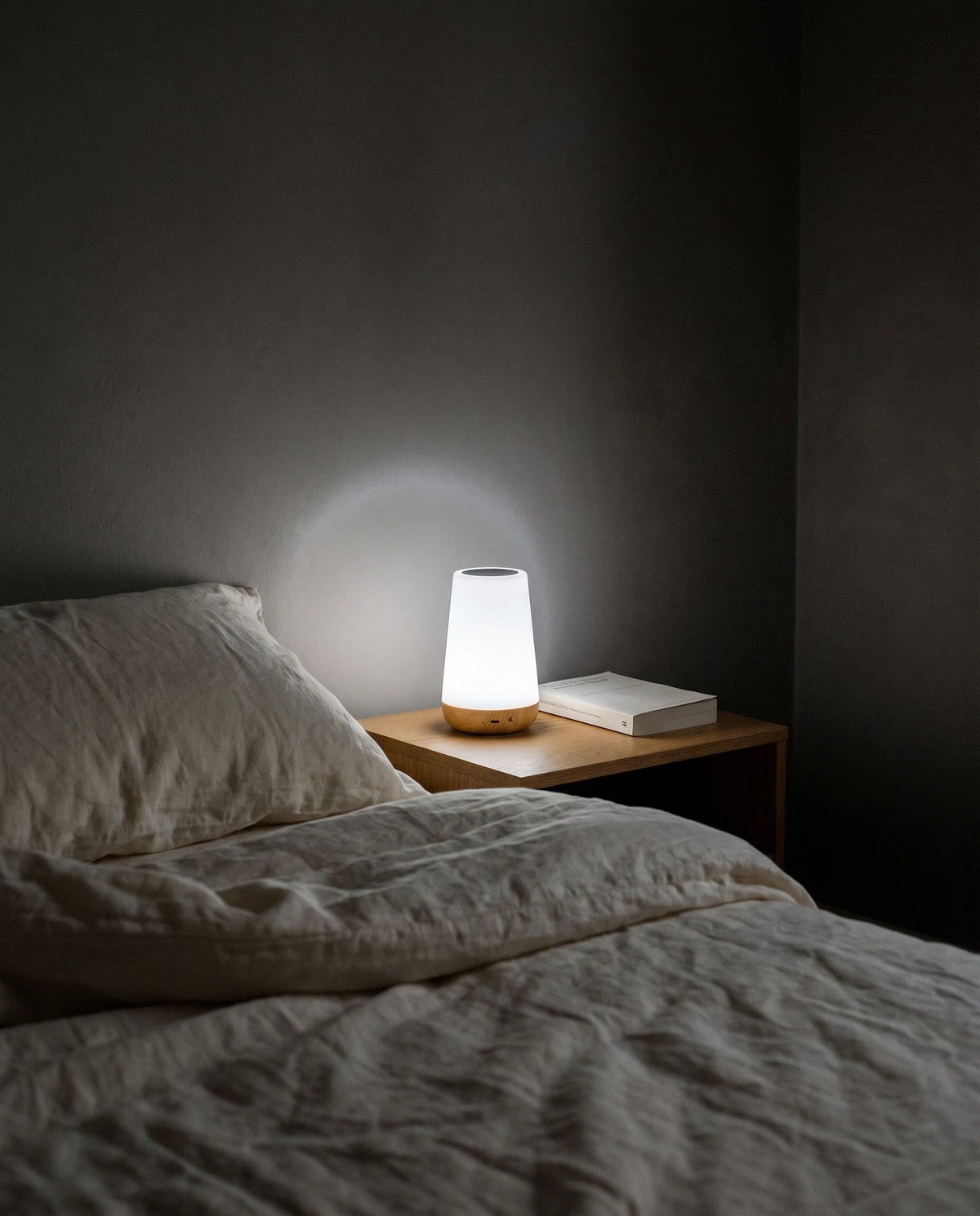 A dark bedroom at night with the lights off: the Lumivano Thimble glows soft white on a small oak bedside table next to a closed paperback, throwing a gentle halo onto the dark grey wall, beside a bed with rumpled ivory linen.