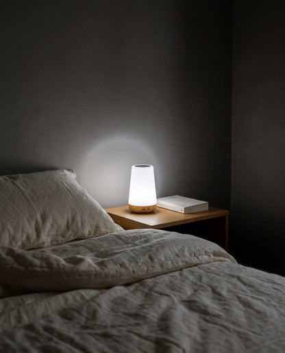 A dark bedroom at night with the lights off: the Lumivano Thimble glows soft white on a small oak bedside table next to a closed paperback, throwing a gentle halo onto the dark grey wall, beside a bed with rumpled ivory linen.