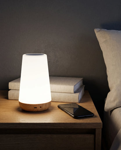 Close view of the Lumivano Thimble glowing soft white on an oak bedside table in the evening, in front of two linen-covered books, with a smartphone lying face down beside it and the edge of a linen pillow on the right. The wood-grain base shows its charging port, pinhole and small slide switch.