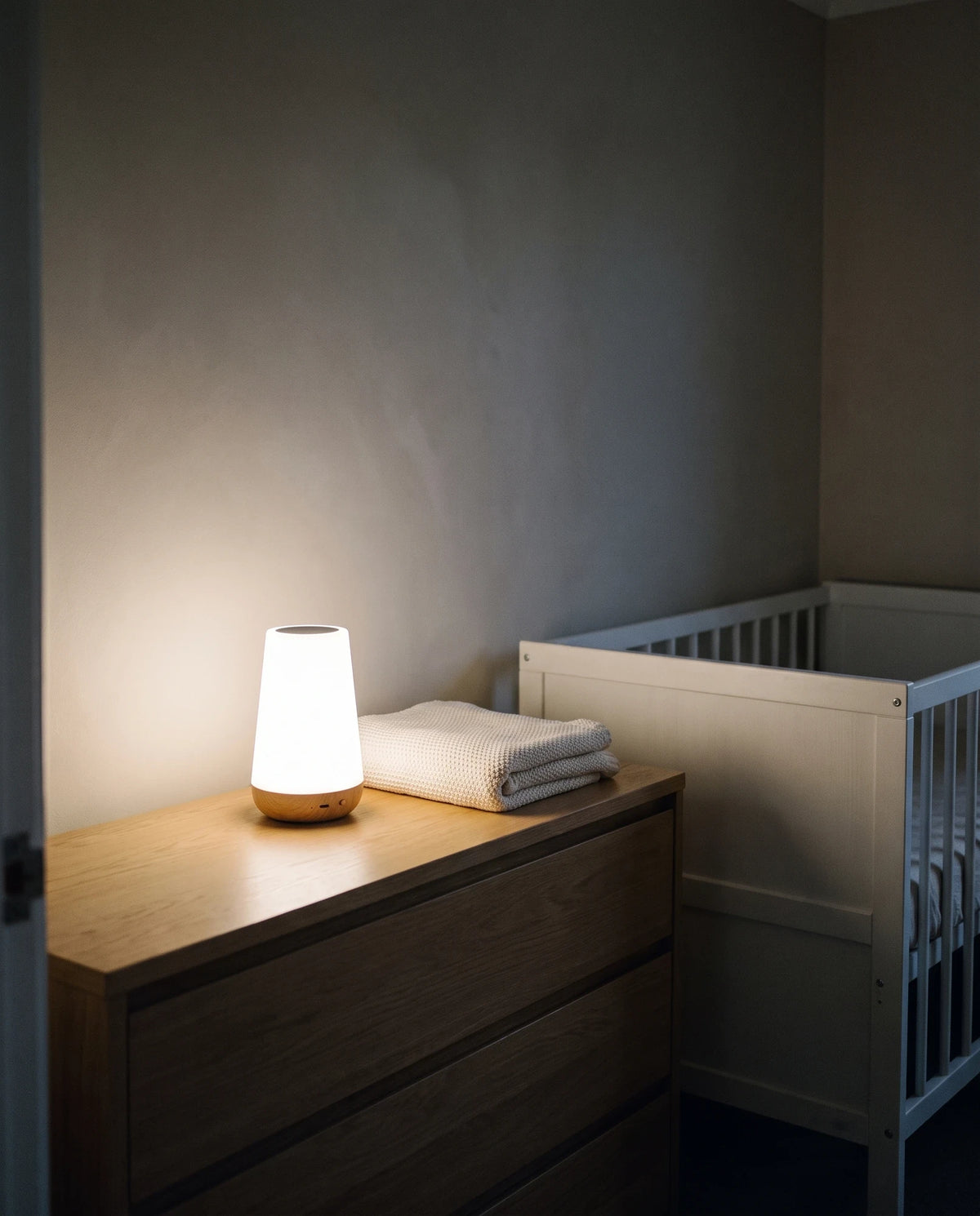 A quiet nursery at night seen from the doorway: the Lumivano Thimble glows soft white on a low oak chest of drawers beside a folded knitted blanket, next to an empty white cot in soft shadow.