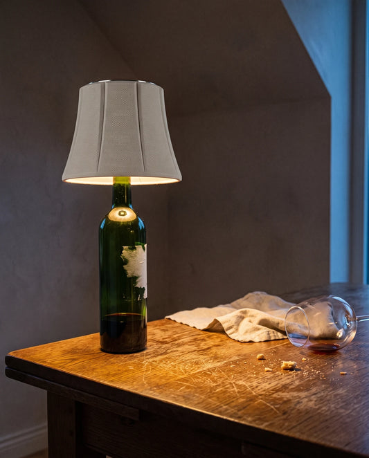 The Lumivano Vintner on the mouth of a green wine bottle with its label peeled off, on the corner of a scratched oak table: the shade throws a warm pool of light down onto the wood, with a crumpled linen napkin, crumbs and a fallen wine glass inside the pool and the plaster wall behind in shadow.