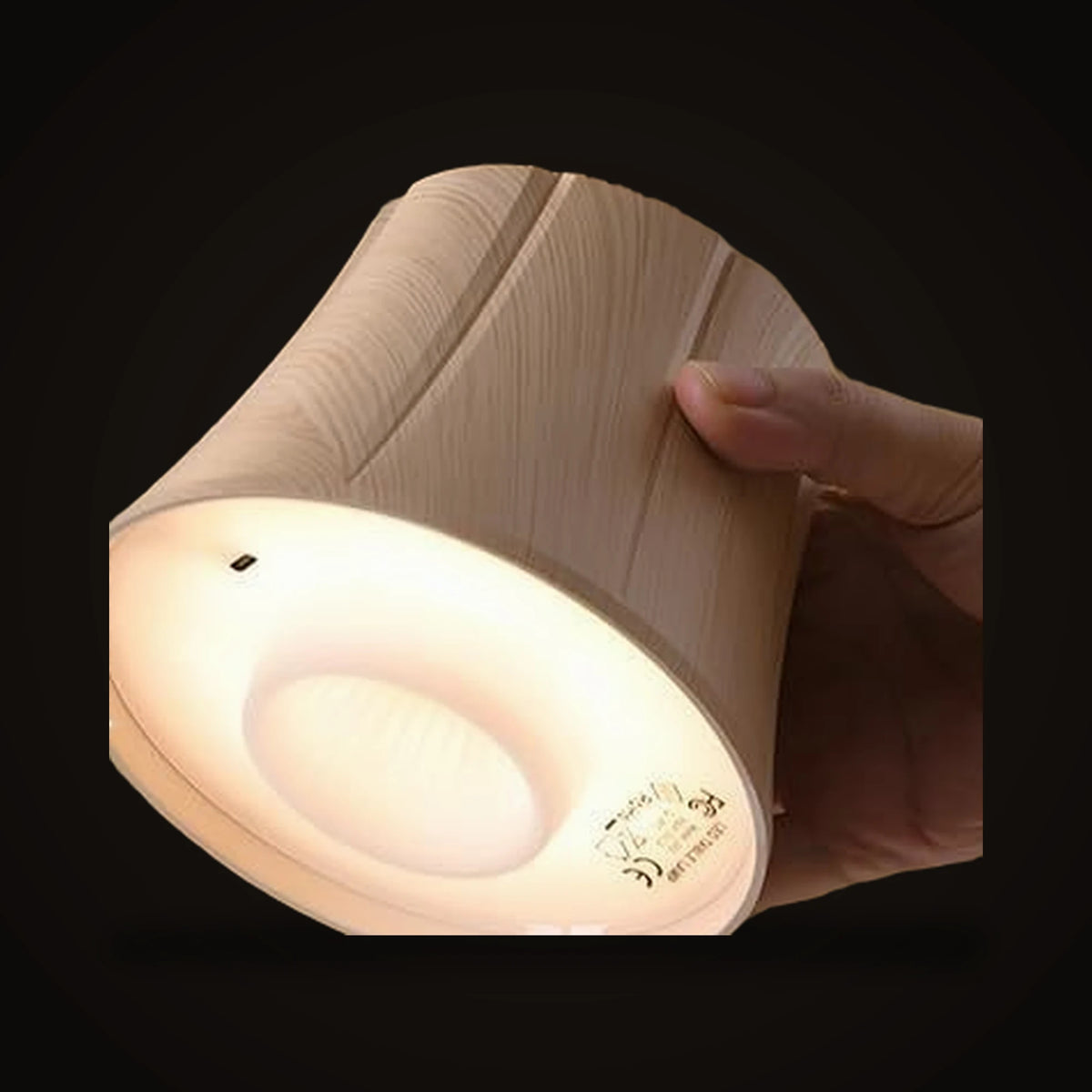 The Lumivano Vintner held in a hand and tilted so its underside shows: the lit face inside the shade, the small charging port on the rim, and the faux wood grain of the outside, on the dark brand ground.
