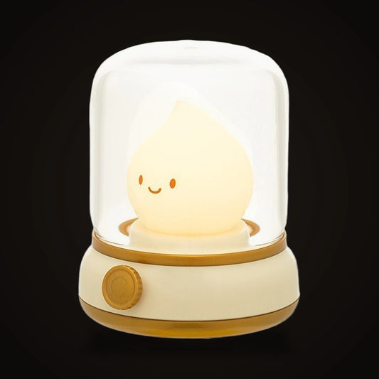The Lumivano Wick on the dark brand ground: a cream silicone flame with two orange dot eyes and a small closed smile, standing under a clear domed cover on an ivory lantern base with two brass rings and a round brass dial.
