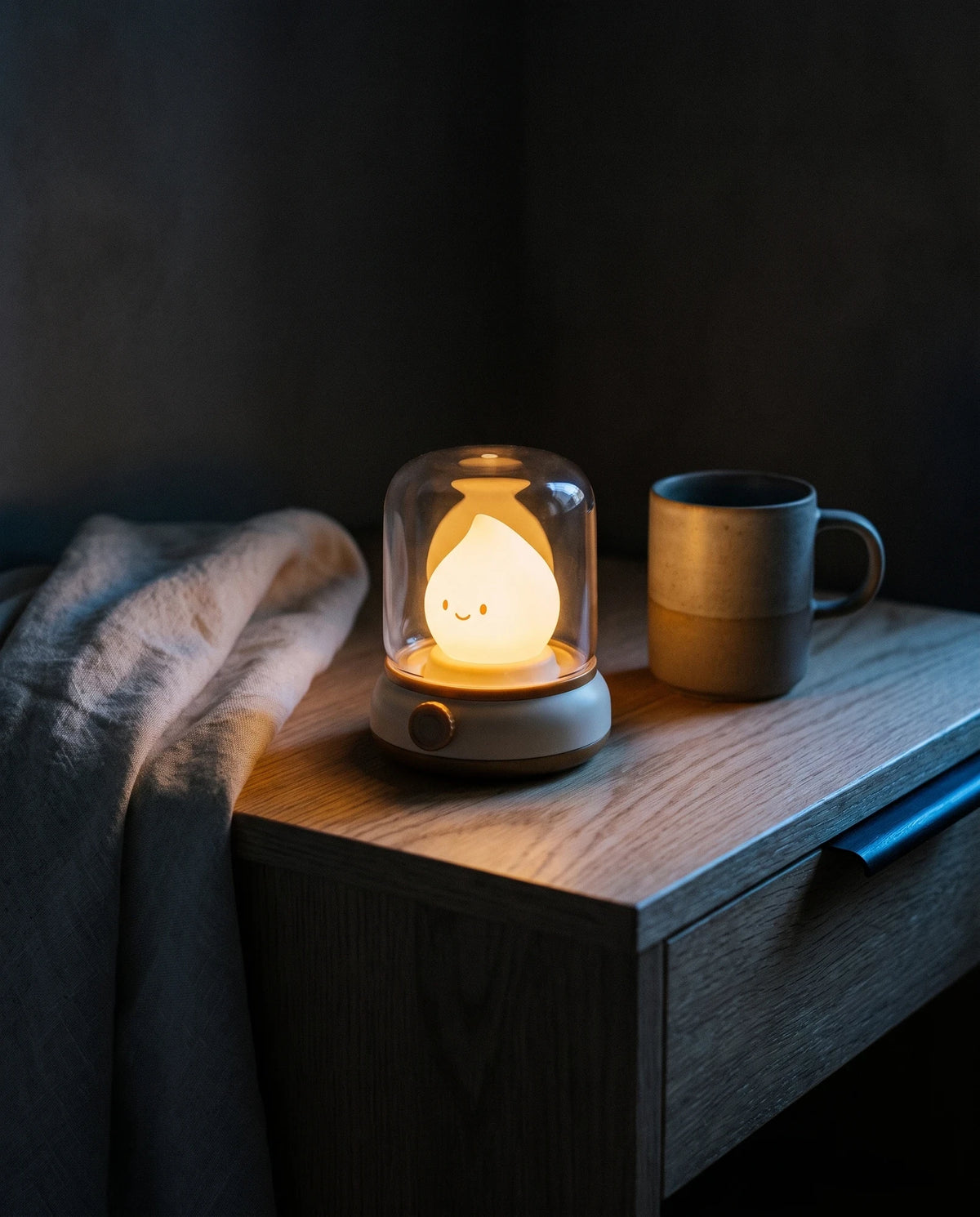 The Lumivano Wick lit on a pale oak bedside table beside a folded ivory linen throw and a matte stoneware cup, throwing warm amber light onto a dark plaster wall, with a narrow cool window glow along the table edge.