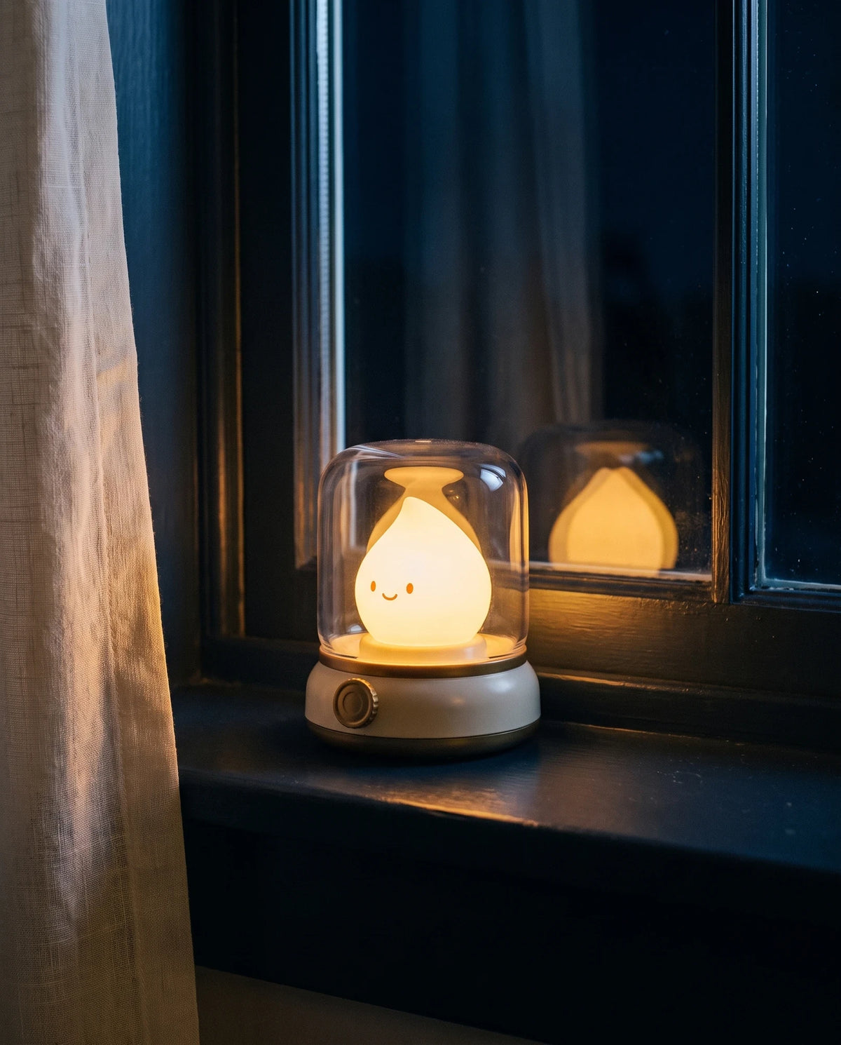 The Lumivano Wick standing alone on a dark painted windowsill at night, its warm flame reflected in the black glass behind it, with a corner of ivory linen curtain lit at the left.