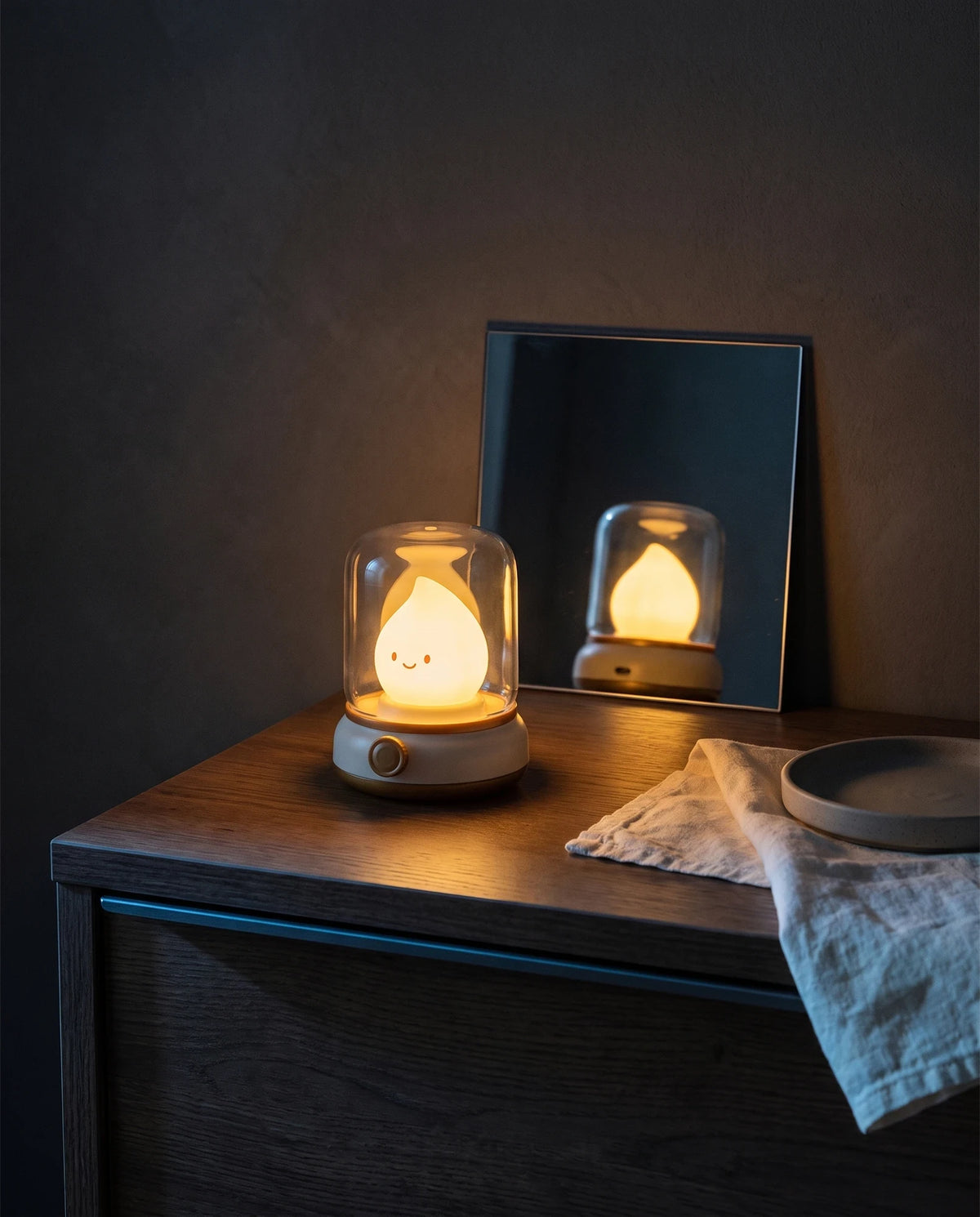 The Lumivano Wick lit on a dark oak dresser with a small mirror leaning on the dark plaster wall behind it, so the back of the lantern and its charging port are visible in the reflection, beside a folded ivory linen cloth and a stoneware dish.
